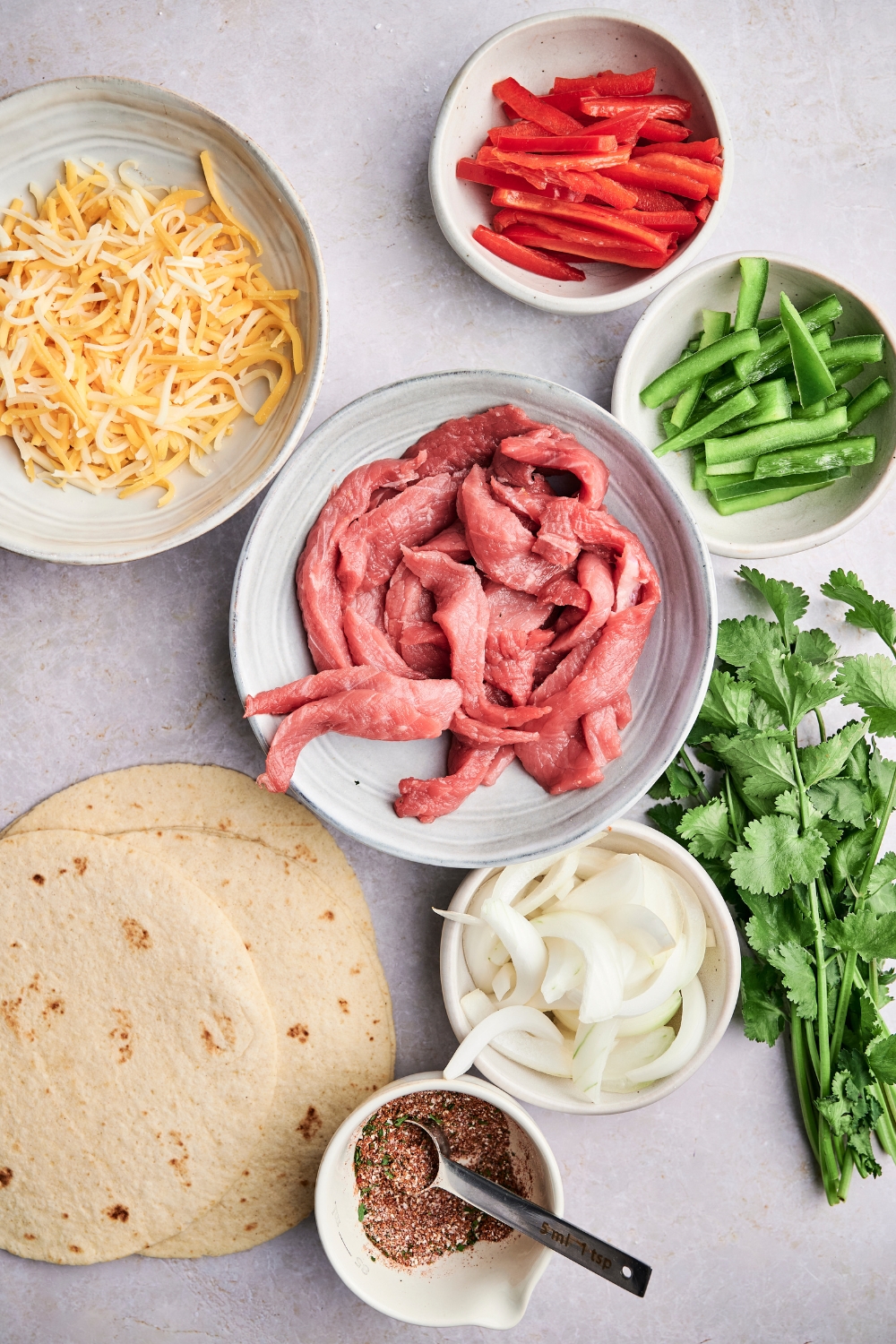 Steak strips, bell peppers, shredded cheese, tortillas, onions, taco seasoning, and fresh cilantro are in separate containers on a counter top.