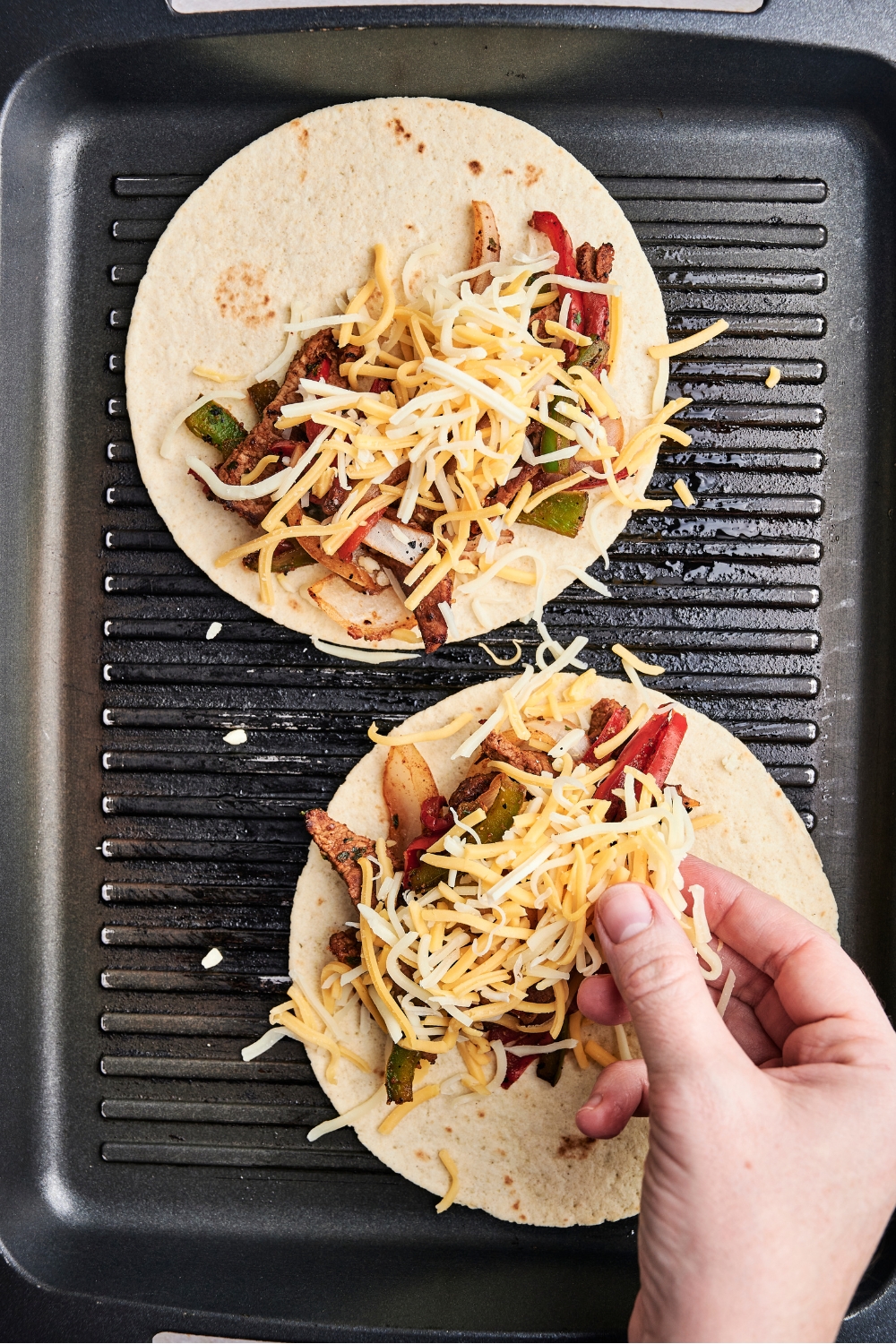 Someone sprinkles shredded cheddar cheese over two open tortillas on a grill pan. The tortillas are also holding steak and vegetables.