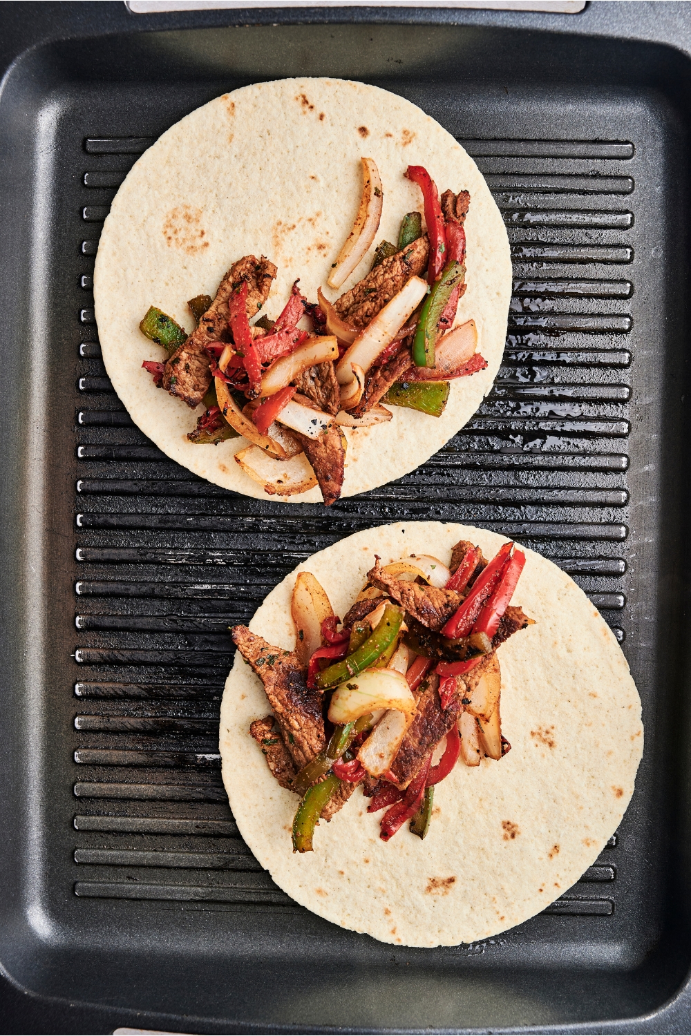 Two tortillas are on a grill tray. There is steak and veggies on each tortilla.
