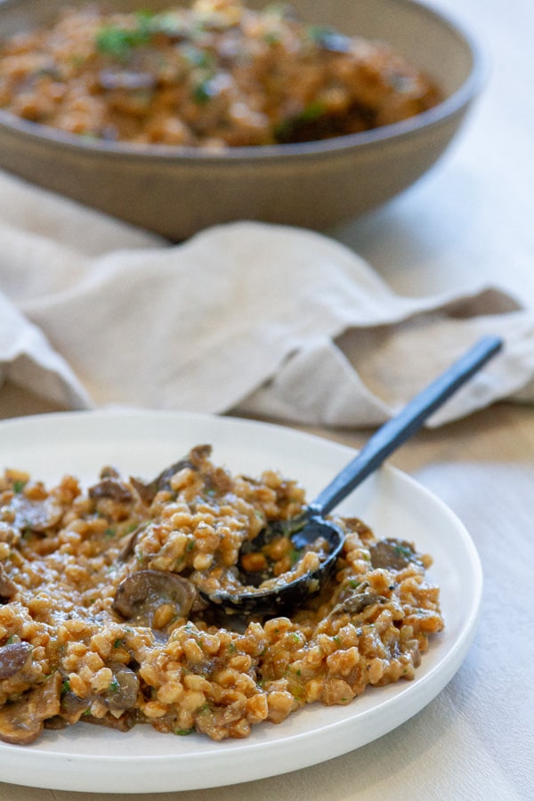 a serving of farro on a white plate with a spoon in front of the serving bowl with a cloth napkin in between.