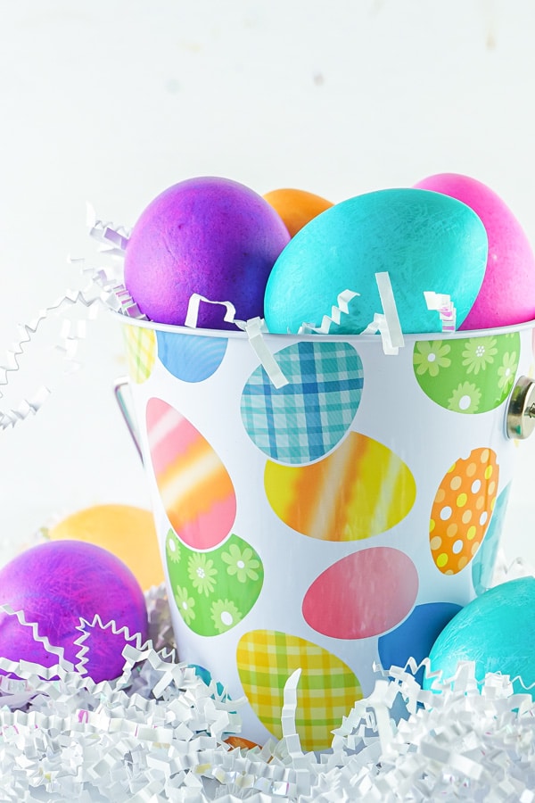 Easter eggs in a tin pail that's painted with Easter decorations nestled in white Easter grass.