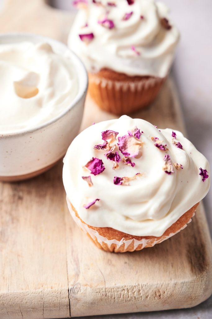 Two cupcakes with creme fraiche frosting and garnished with edible flower pieces sitting on a wooden board.