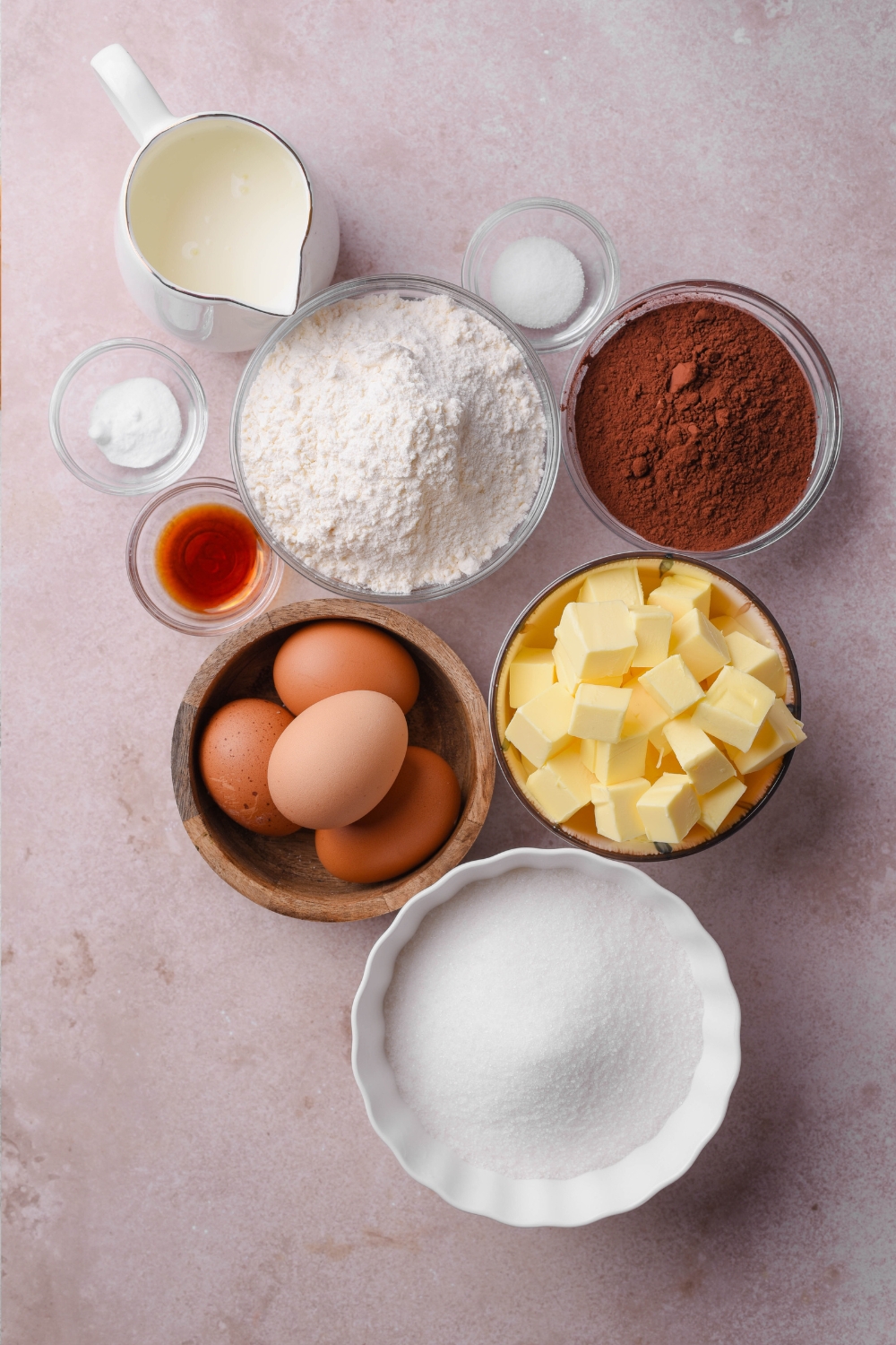 Buttermilk, flour, salt, baking powder, cocoa powder, cubed butter, eggs, vanilla, and white sugar are all in separate bowls on a counter top.