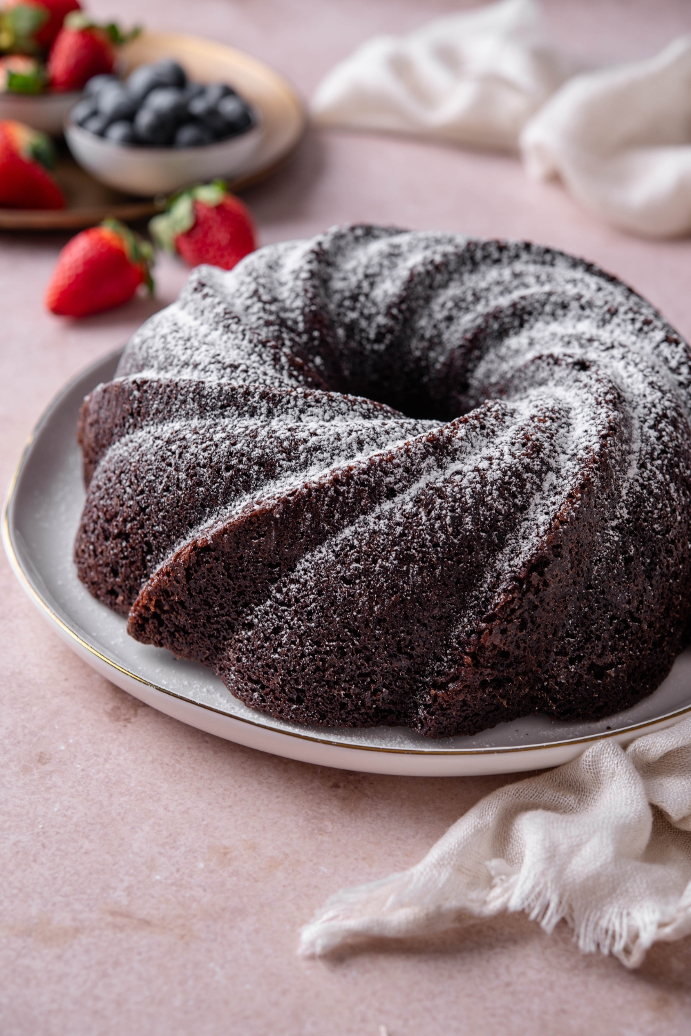 A whole chocolate pound cake is sitting on a white serving plate. Fresh berries are near by.