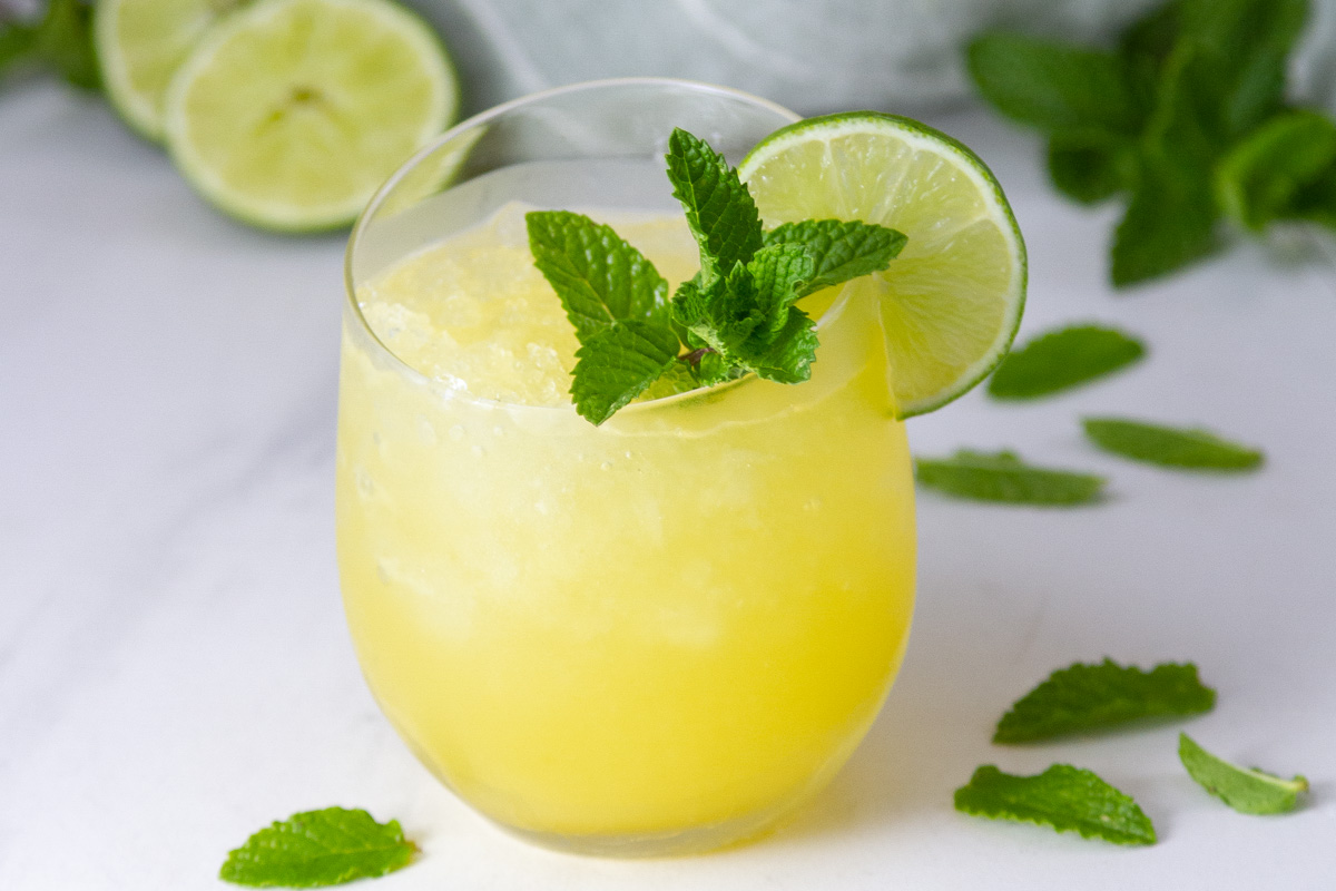 a glass filled with pineapple drink garnished with lime wheel and mint.