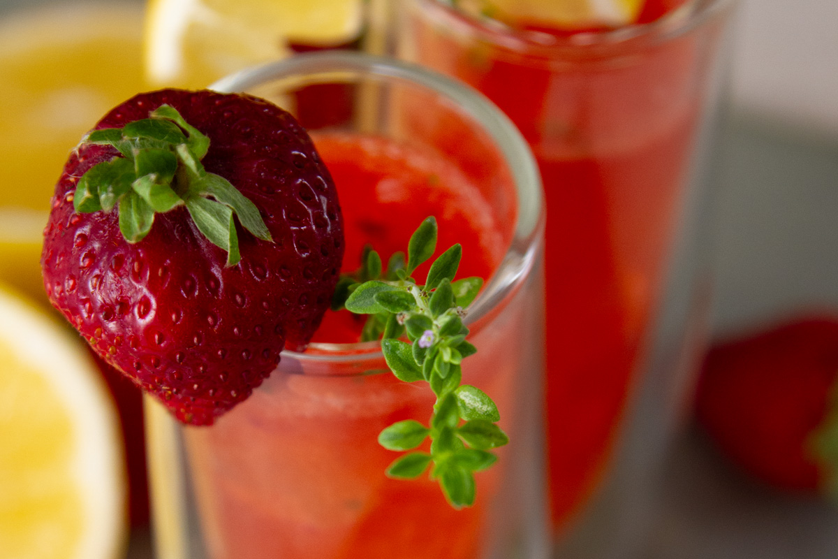a tall glass of strwaberry drink garnishes with a strawberry and some fresh thyme.
